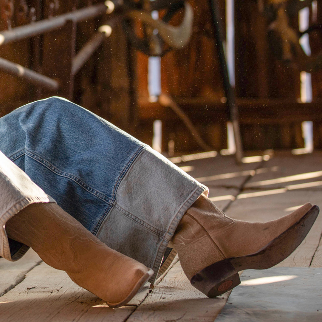 Person wearing brown cowboy boots and blue jeans on a wooden floor.