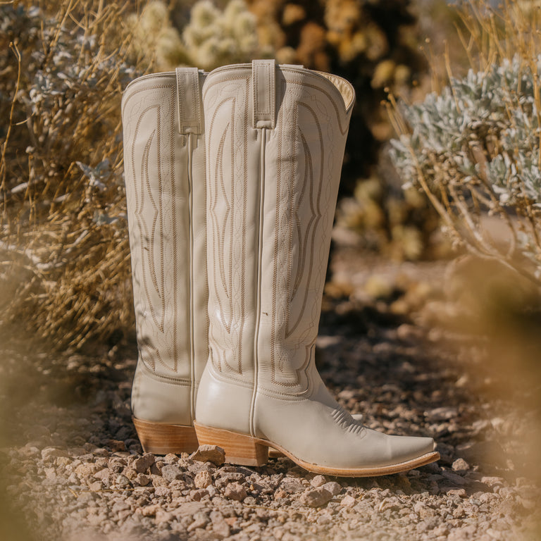 Pair of light-colored cowboy boots on a rocky ground with desert plants in the background
