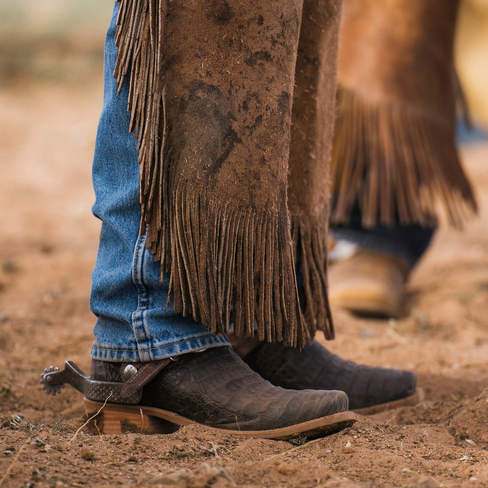 Man standing in dirt wearing cowboy boots, blue jeans and brown suede chaps with blurred background