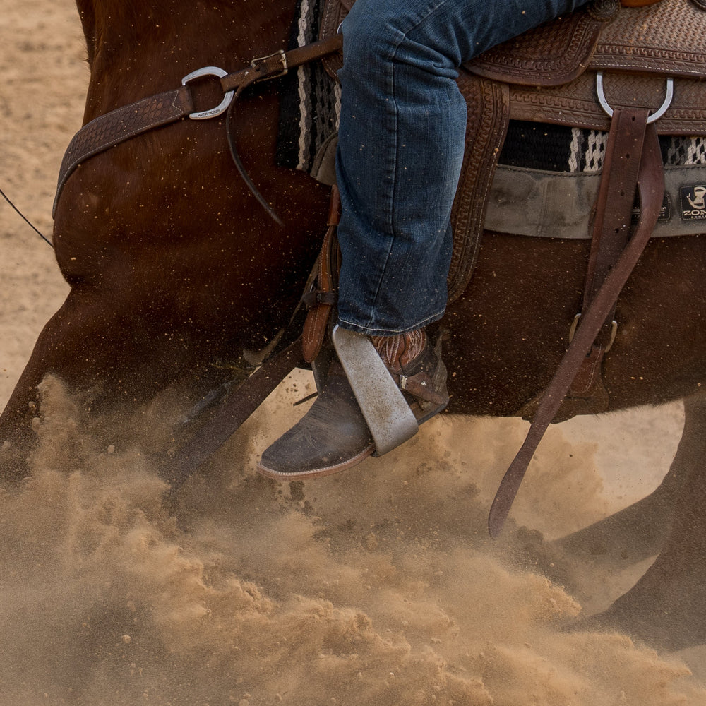 Photo of man on horse focusing on the stirrup and cowboy boot with dust kicking up in the background
