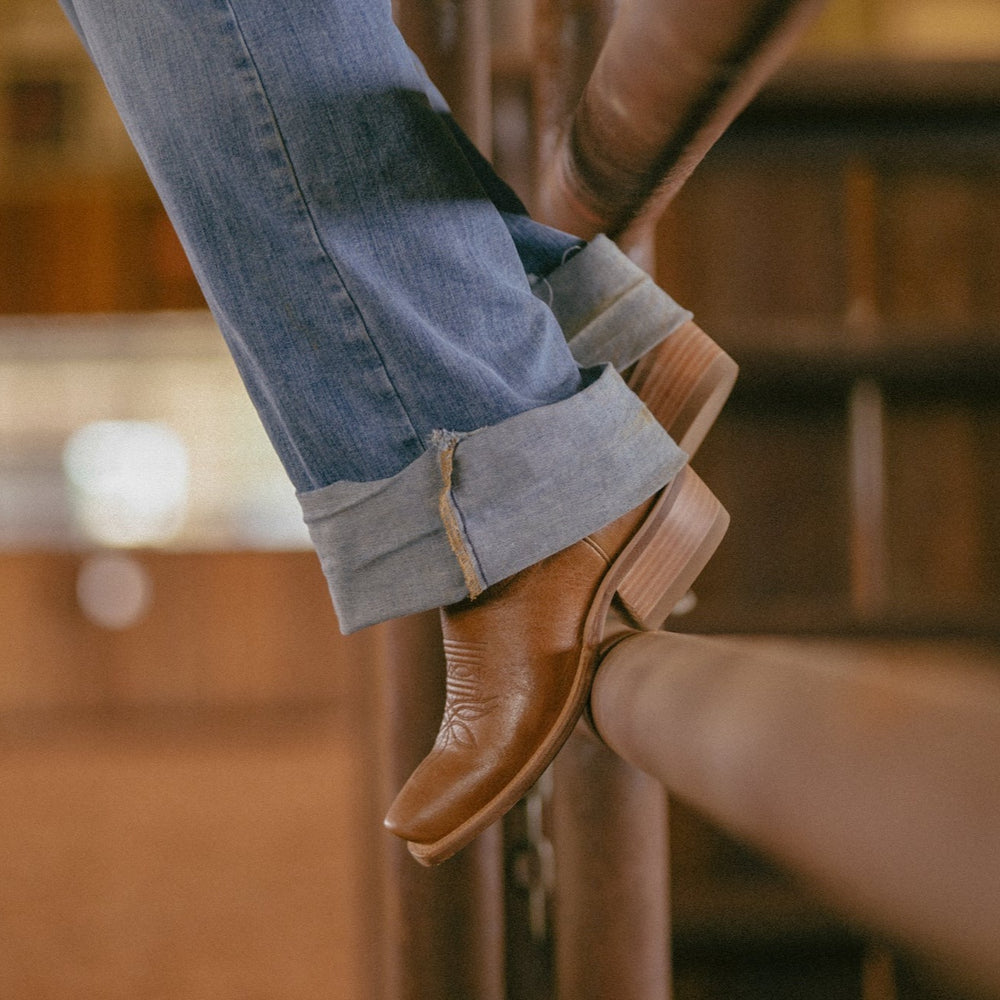 Close up crop of a person wearing blue jeans and brown cowboy boots sitting on a fence