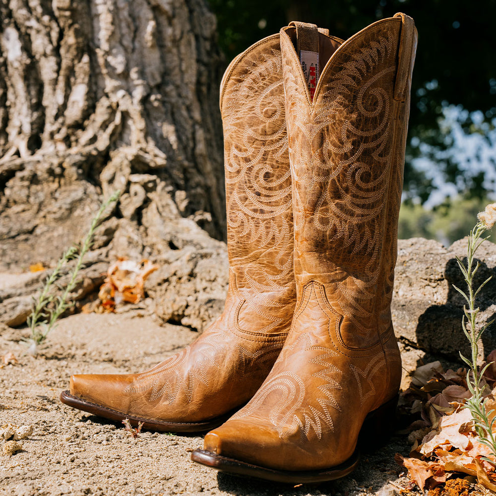 Pair of light brown snip toe cowboy boots with allover light tan stitching sitting on a dirt in front of a tree