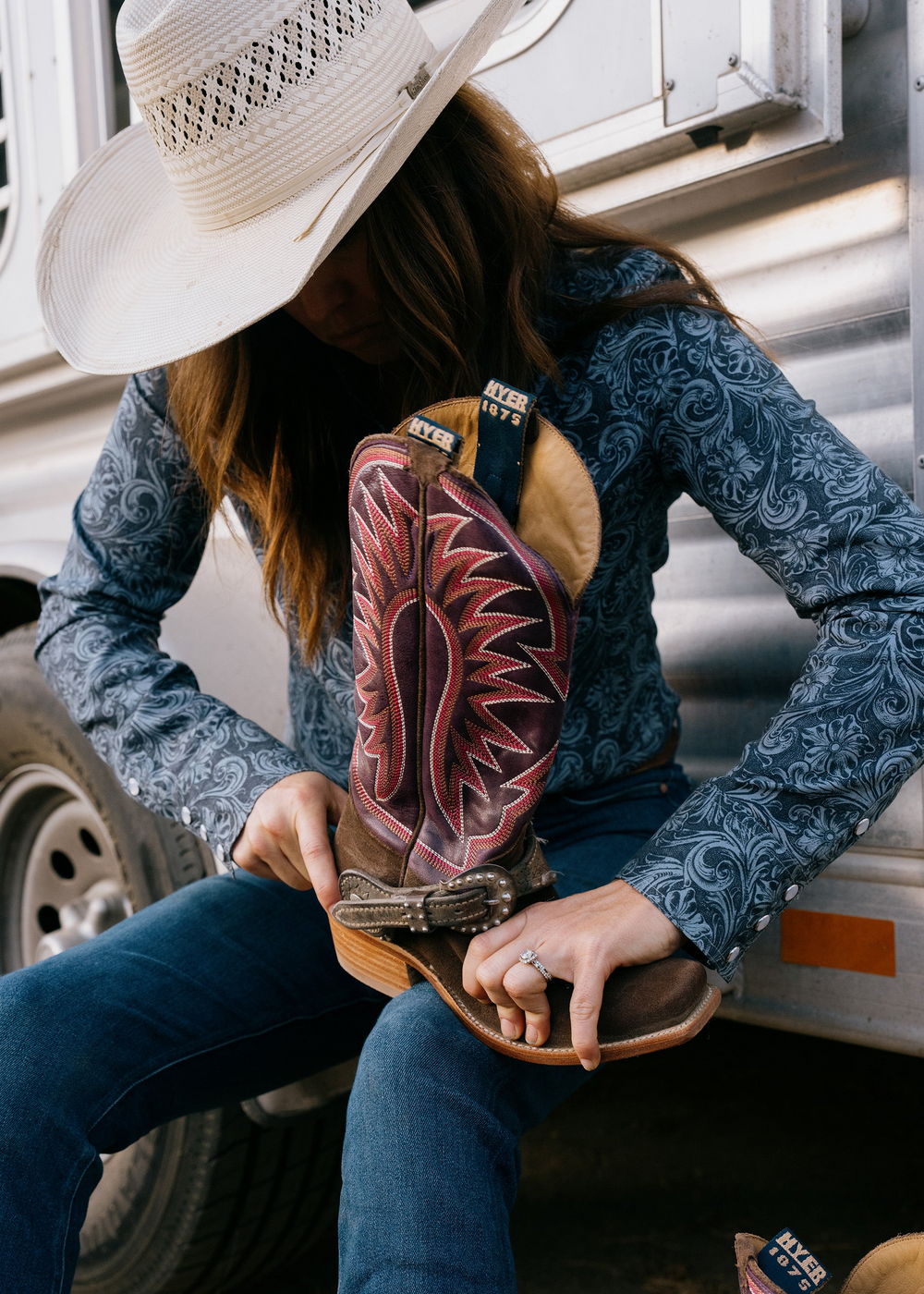 Lifestyle shot of cowgirl wearing white cowboy hat, blue detailed shirt, and blue jeans holding a brown cowgirl boot with a spur attached sitting on the end of a trailer