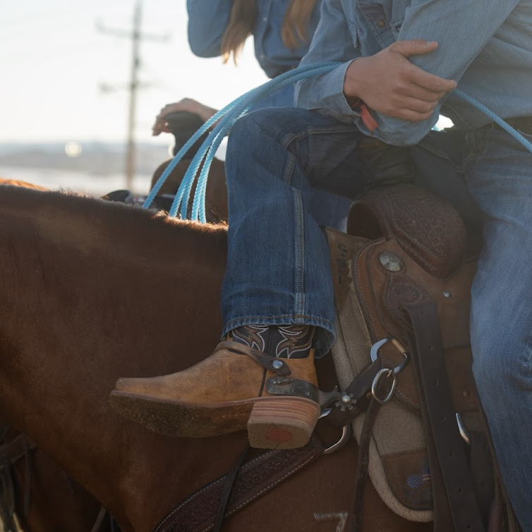 Person sitting on a horse wearing cowboy boots and blue jeans, with a blurred background.