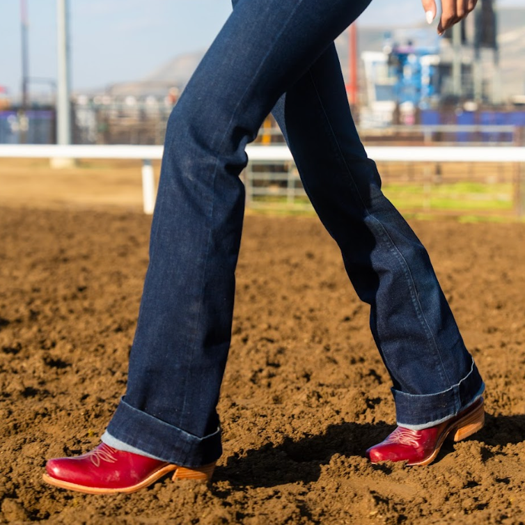 Person wearing blue jeans and red cowboy boots on a dirt field.