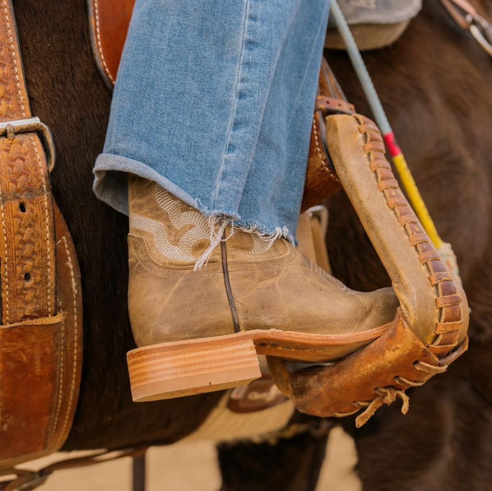 Close-up of a person wearing brown leather boots and blue jeans on a horse, with a saddle and bridle in the background.