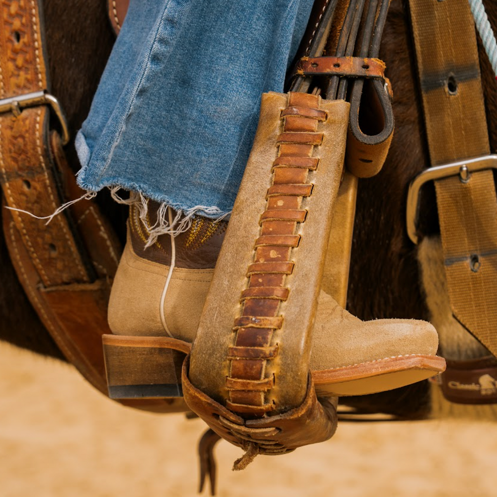 Close-up of beige cowboy boots with brown leather details in stirrup, worn jeans, and a saddle in the background.